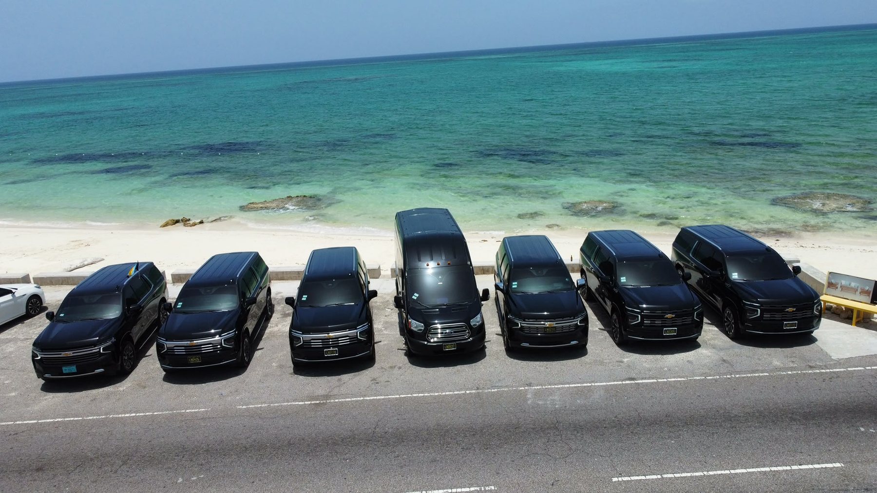 Seven black Johnson Executive vehicles parked in a row facing the turquoise water at Cable Beach, Nassau